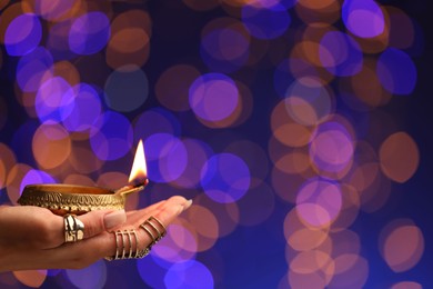 Photo of Diwali celebration. Woman holding lit diya lamp on color background with blurred lights, closeup. Space for text