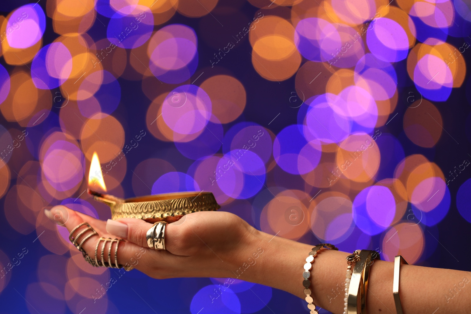 Photo of Diwali celebration. Woman holding lit diya lamp on color background with blurred lights, closeup