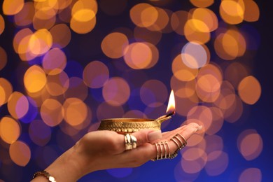 Photo of Diwali celebration. Woman holding lit diya lamp on color background with blurred lights, closeup