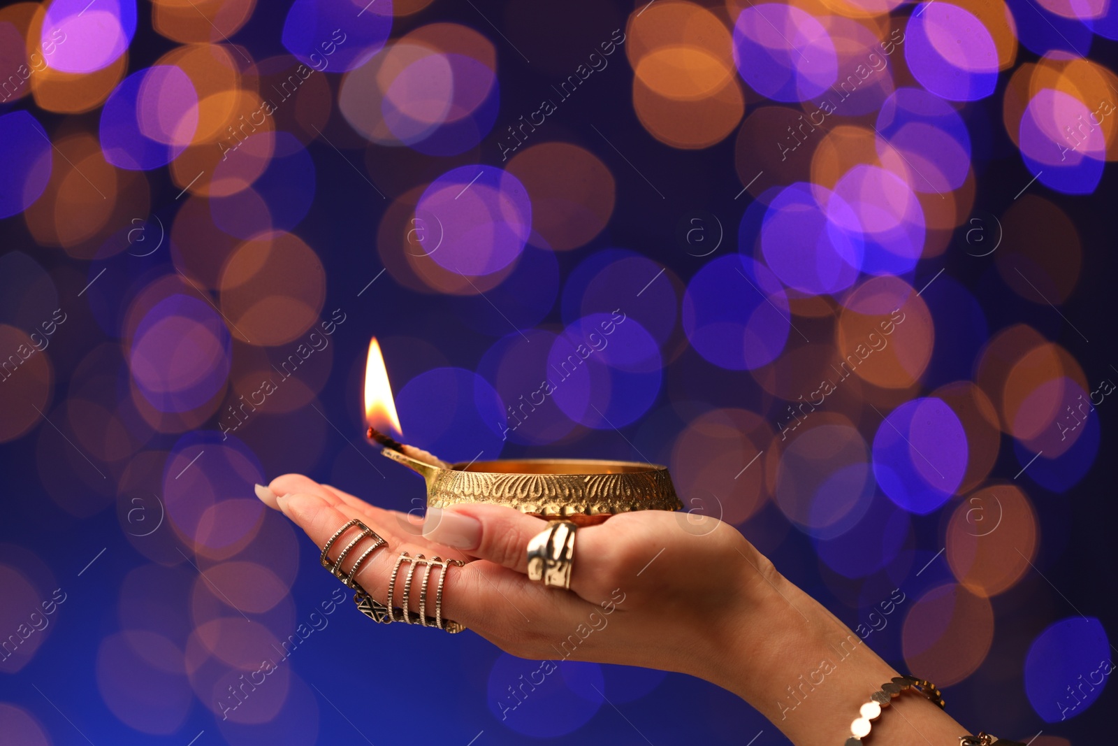 Photo of Diwali celebration. Woman holding lit diya lamp on color background with blurred lights, closeup