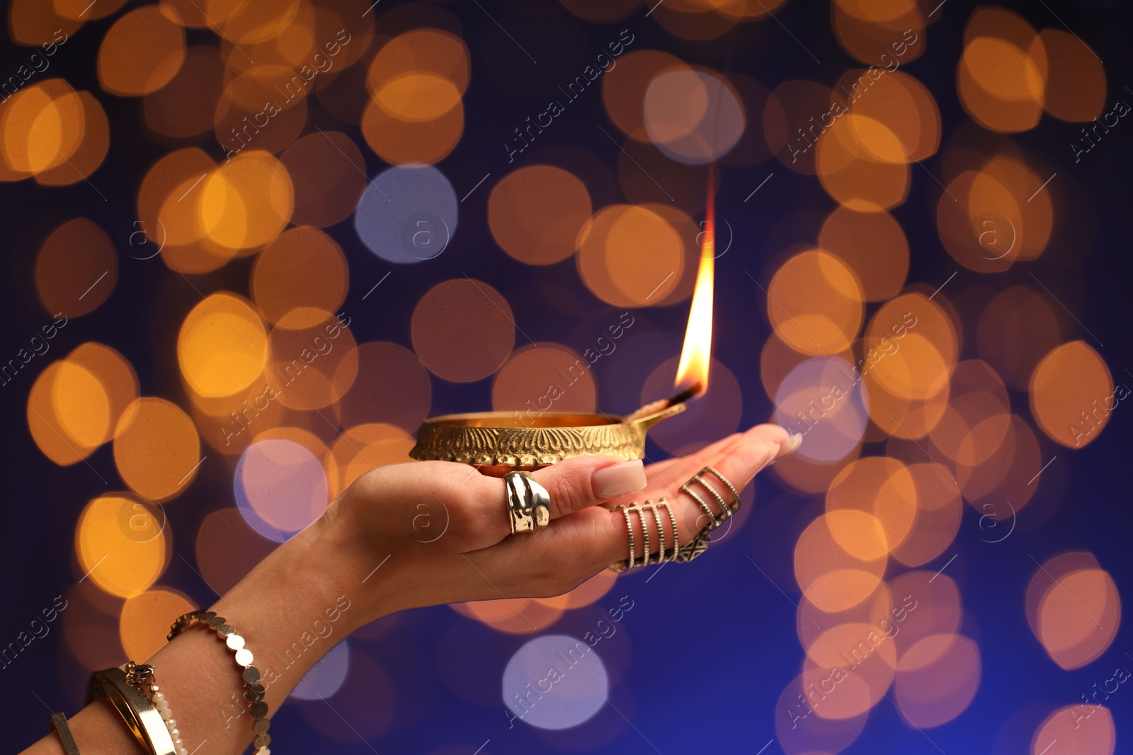 Photo of Diwali celebration. Woman holding lit diya lamp on color background with blurred lights, closeup