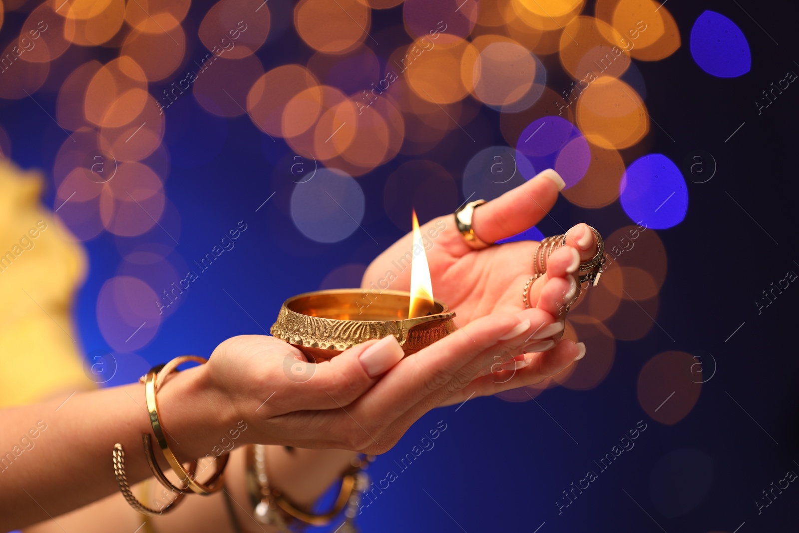 Photo of Diwali celebration. Woman holding lit diya lamp on color background with blurred lights, closeup