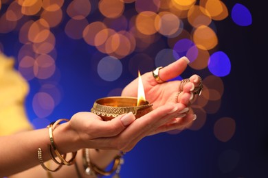 Diwali celebration. Woman holding lit diya lamp on color background with blurred lights, closeup