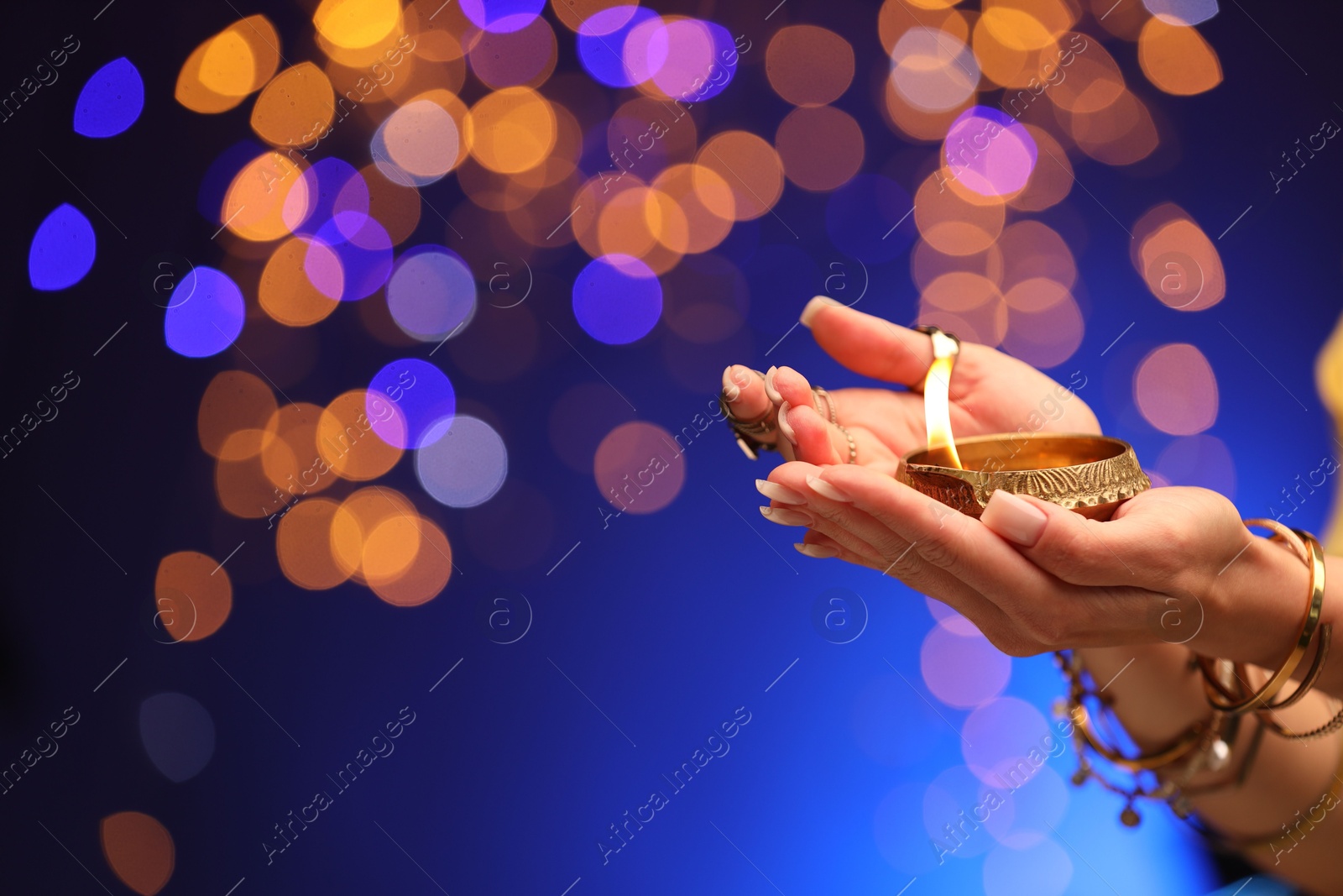 Photo of Diwali celebration. Woman holding lit diya lamp on blue background with blurred lights, closeup. Space for text