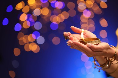 Photo of Diwali celebration. Woman holding lit diya lamp on blue background with blurred lights, closeup. Space for text