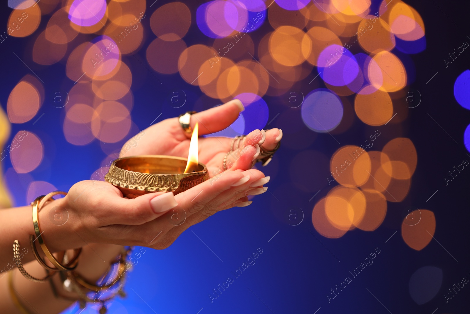 Photo of Diwali celebration. Woman holding lit diya lamp on blue background with blurred lights, closeup