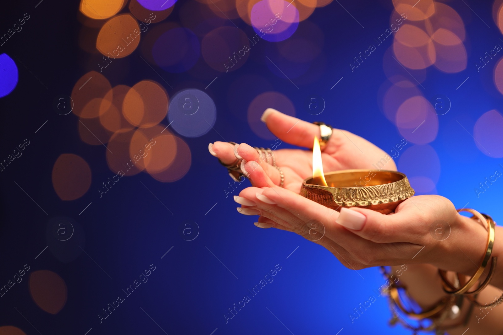 Photo of Diwali celebration. Woman holding lit diya lamp on blue background with blurred lights, closeup