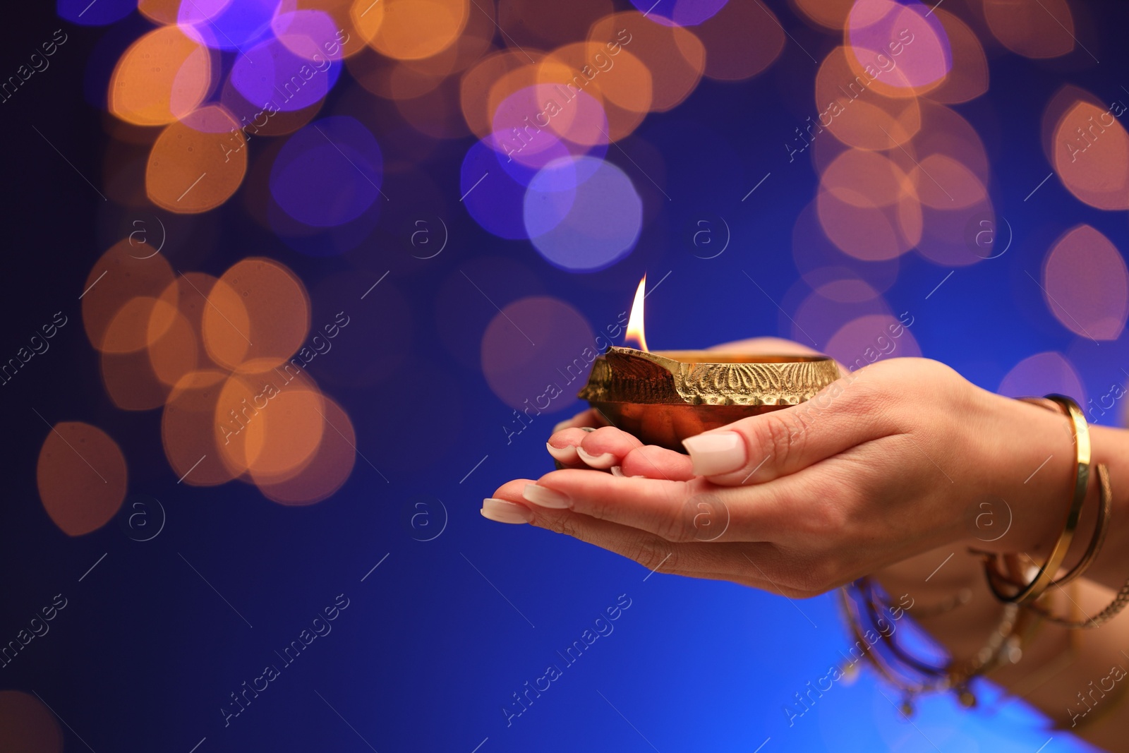 Photo of Diwali celebration. Woman holding lit diya lamp on blue background with blurred lights, closeup