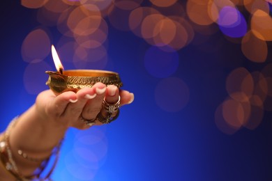 Photo of Diwali celebration. Woman holding lit diya lamp on blue background with blurred lights, closeup. Space for text