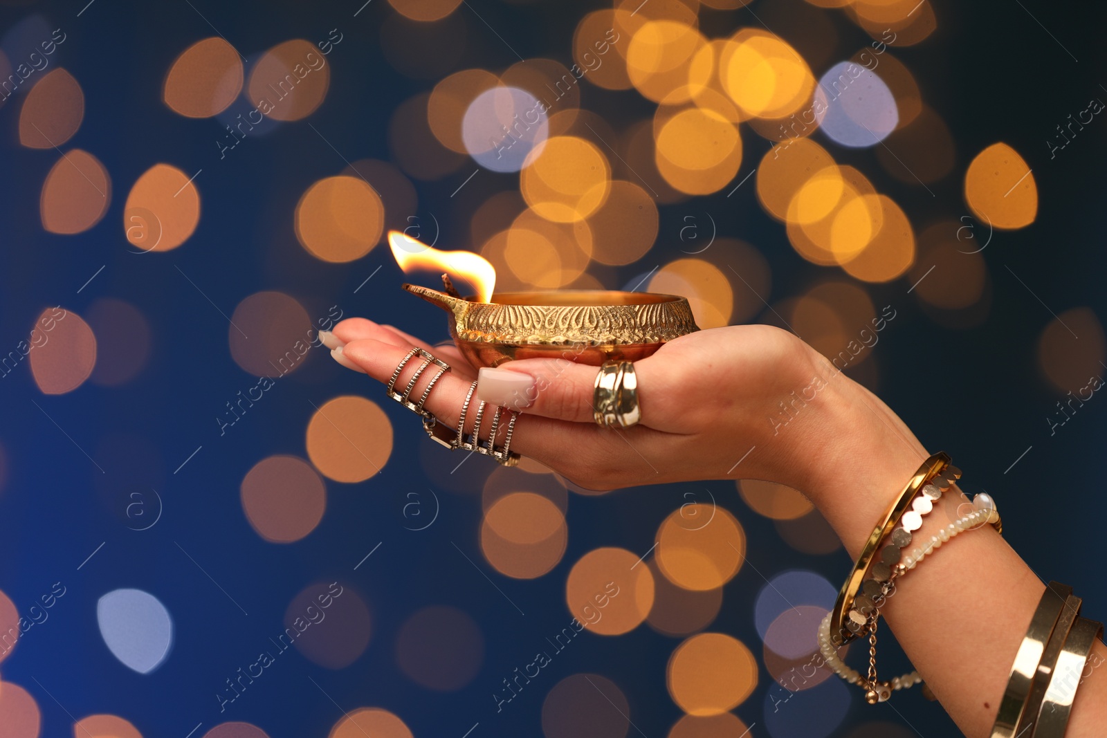 Photo of Diwali celebration. Woman holding lit diya lamp on blue background with blurred lights, closeup