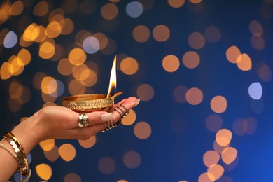 Diwali celebration. Woman holding lit diya lamp on blue background with blurred lights, closeup. Space for text