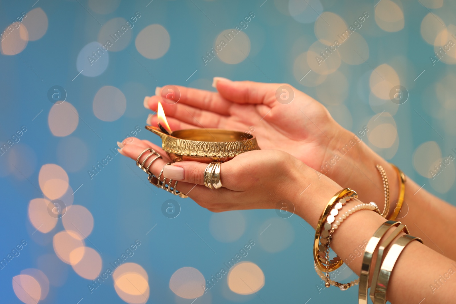 Photo of Diwali celebration. Woman holding lit diya lamp on light blue background with blurred lights, closeup