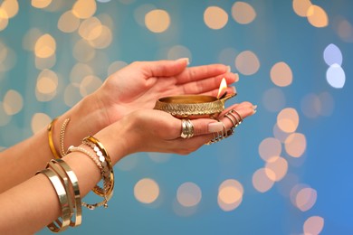 Photo of Diwali celebration. Woman holding lit diya lamp on light blue background with blurred lights, closeup