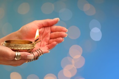 Photo of Diwali celebration. Woman holding lit diya lamp on light blue background with blurred lights, closeup. Space for text