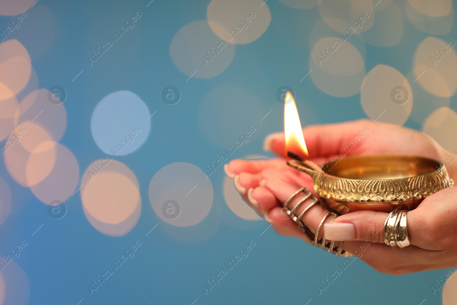 Photo of Diwali celebration. Woman holding lit diya lamp on light blue background with blurred lights, closeup. Space for text