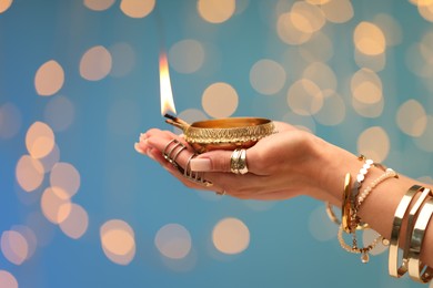 Photo of Diwali celebration. Woman holding lit diya lamp on light blue background with blurred lights, closeup