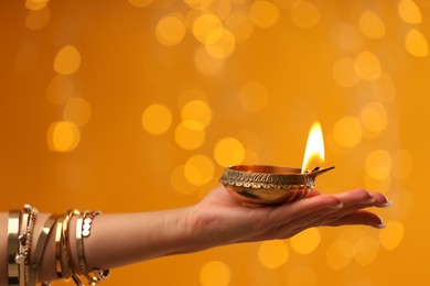 Photo of Diwali celebration. Woman holding lit diya lamp on orange background with blurred lights, closeup