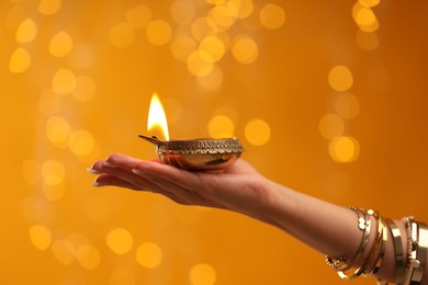 Photo of Diwali celebration. Woman holding lit diya lamp on orange background with blurred lights, closeup