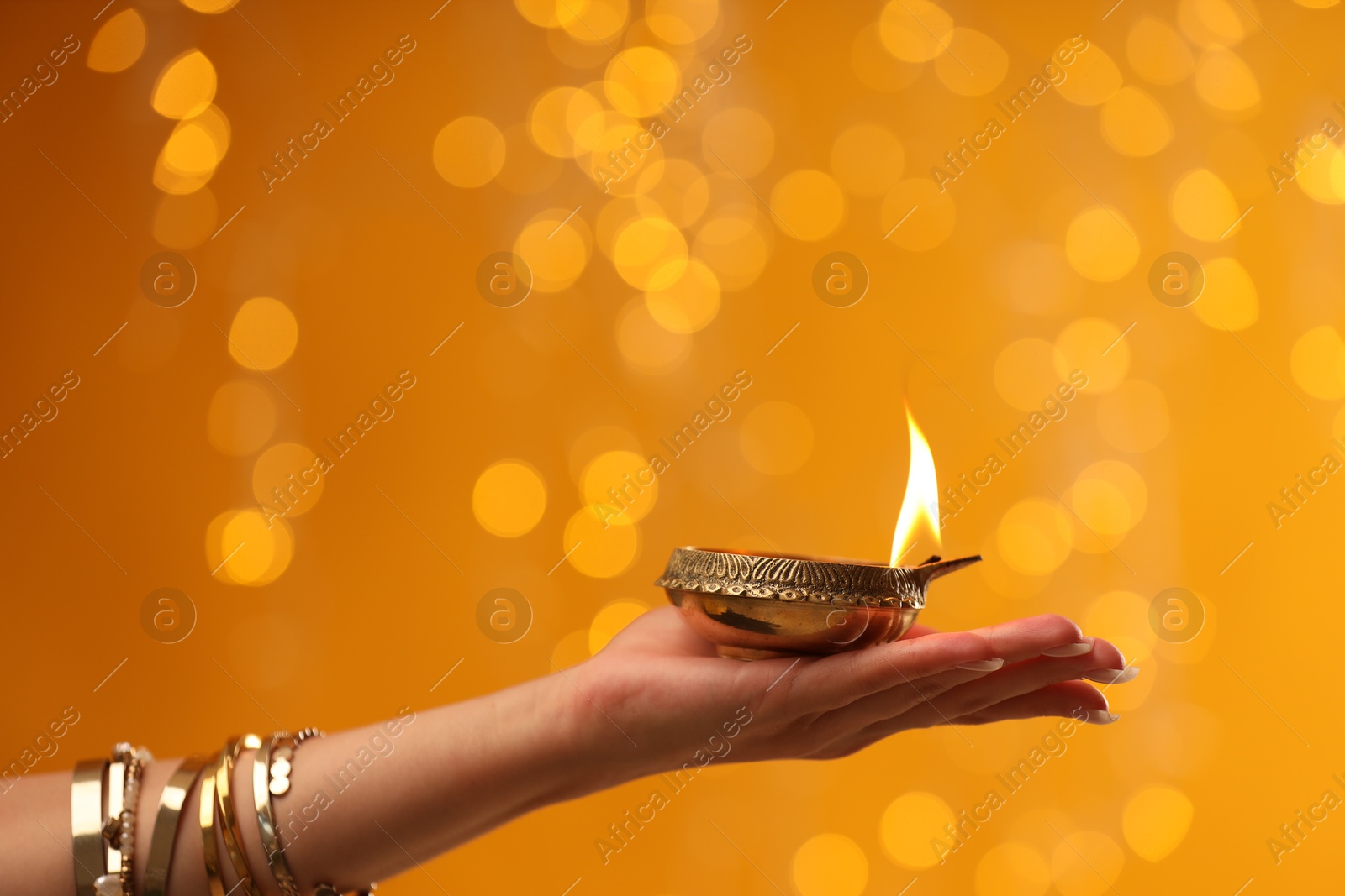 Photo of Diwali celebration. Woman holding lit diya lamp on orange background with blurred lights, closeup