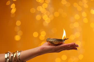 Diwali celebration. Woman holding lit diya lamp on orange background with blurred lights, closeup