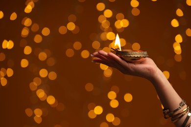 Photo of Diwali celebration. Woman holding lit diya lamp on brown background with blurred lights, closeup. Space for text