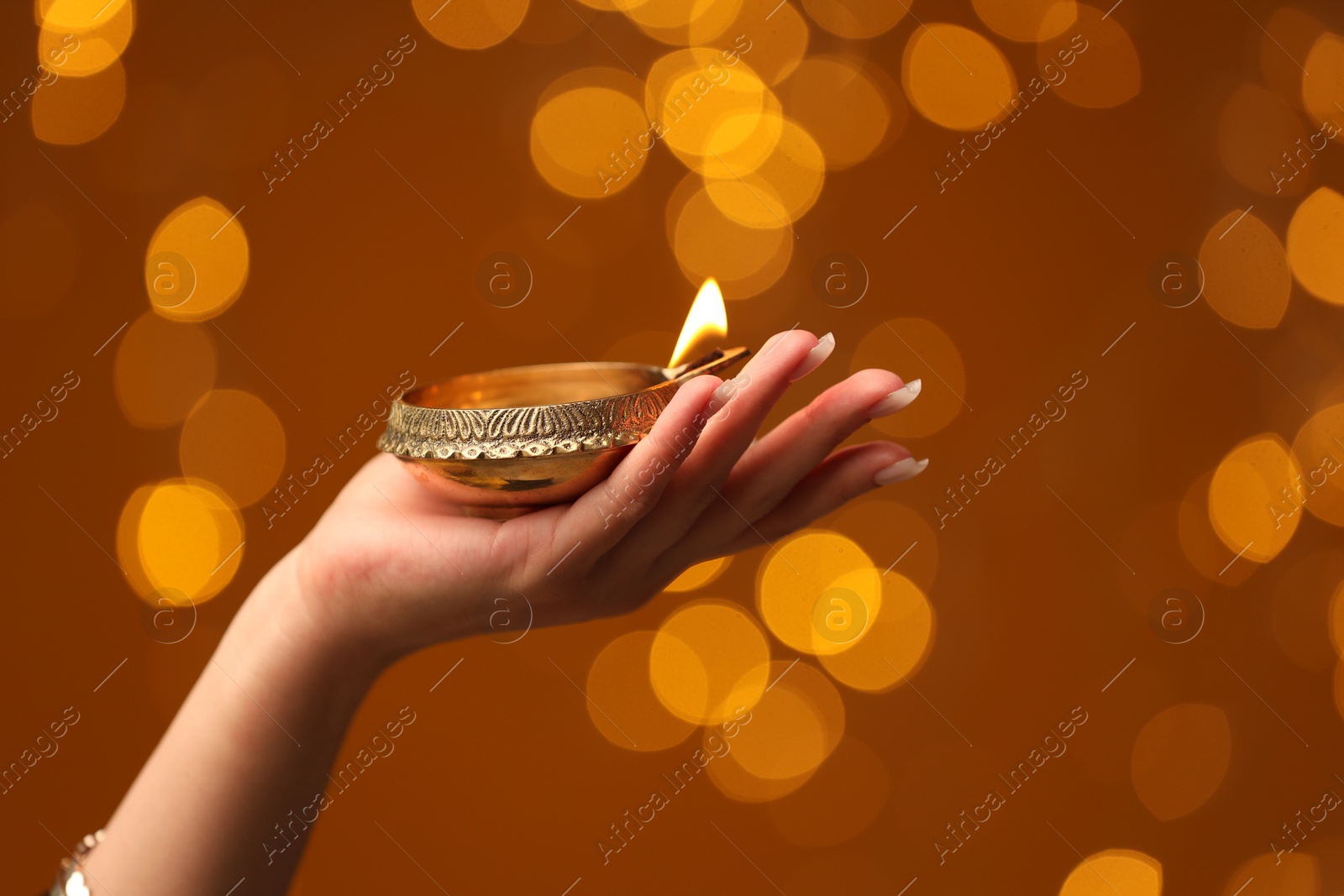 Photo of Diwali celebration. Woman holding lit diya lamp on brown background with blurred lights, closeup