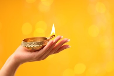 Photo of Diwali celebration. Woman holding lit diya lamp on yellow background with blurred lights, closeup. Space for text