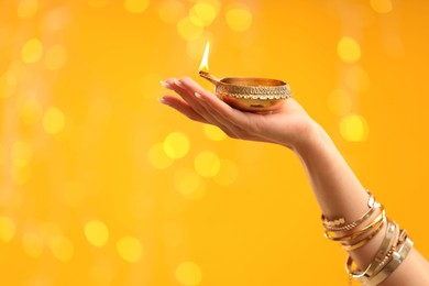 Diwali celebration. Woman holding lit diya lamp on yellow background with blurred lights, closeup. Space for text