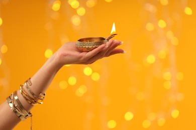 Photo of Diwali celebration. Woman holding lit diya lamp on orange background with blurred lights, closeup. Space for text