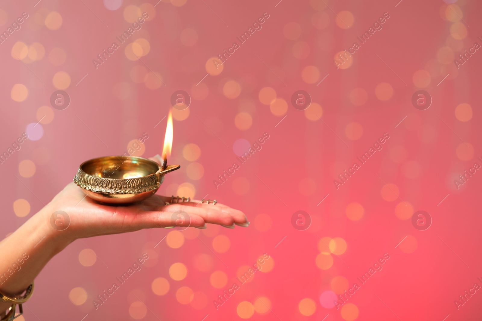 Photo of Diwali celebration. Woman holding lit diya lamp on color background with blurred lights, closeup. Space for text