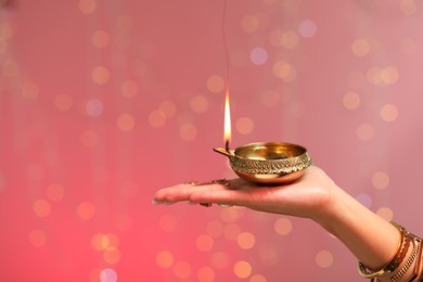 Diwali celebration. Woman holding lit diya lamp on color background with blurred lights, closeup. Space for text