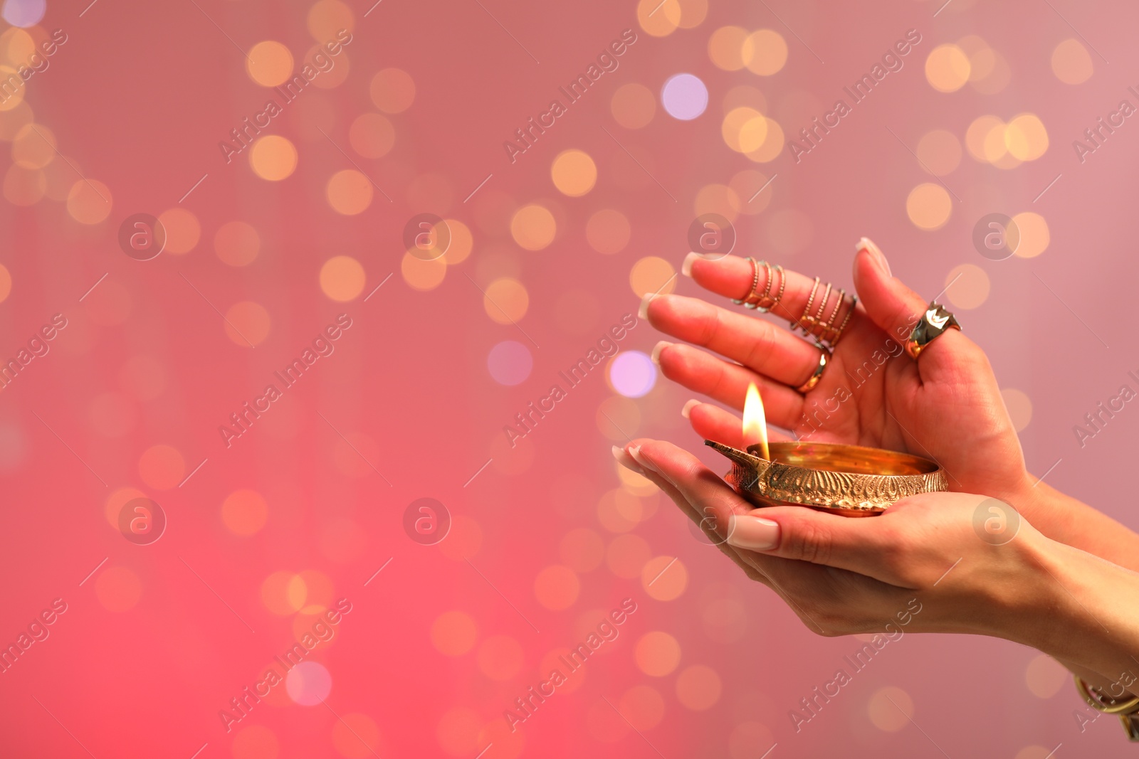 Photo of Diwali celebration. Woman holding lit diya lamp on color background with blurred lights, closeup. Space for text