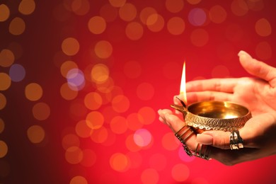 Photo of Diwali celebration. Woman holding lit diya lamp on red background with blurred lights, closeup. Space for text