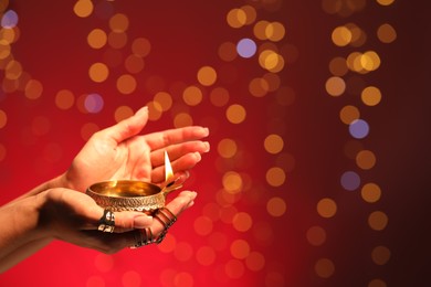 Photo of Diwali celebration. Woman holding lit diya lamp on red background with blurred lights, closeup. Space for text