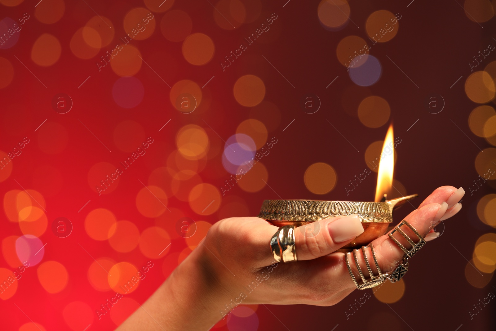 Photo of Diwali celebration. Woman holding lit diya lamp on red background with blurred lights, closeup. Space for text