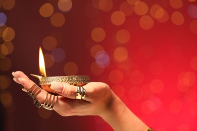 Photo of Diwali celebration. Woman holding lit diya lamp on red background with blurred lights, closeup. Space for text