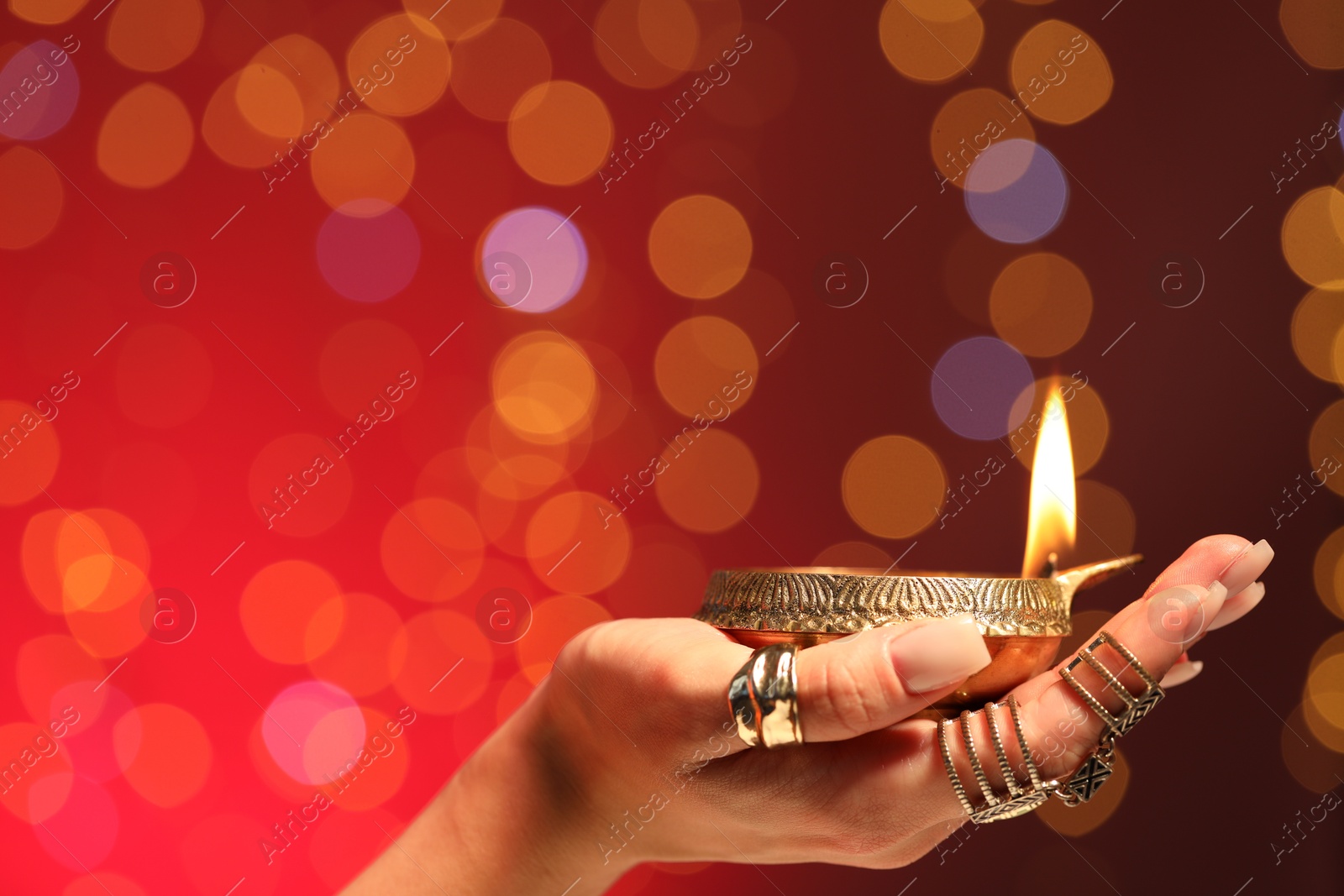 Photo of Diwali celebration. Woman holding lit diya lamp on red background with blurred lights, closeup. Space for text