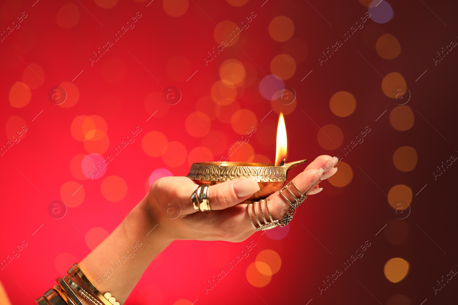 Photo of Diwali celebration. Woman holding lit diya lamp on red background with blurred lights, closeup