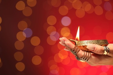 Photo of Diwali celebration. Woman holding lit diya lamp on red background with blurred lights, closeup. Space for text