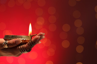 Photo of Diwali celebration. Woman holding lit diya lamp on red background with blurred lights, closeup. Space for text