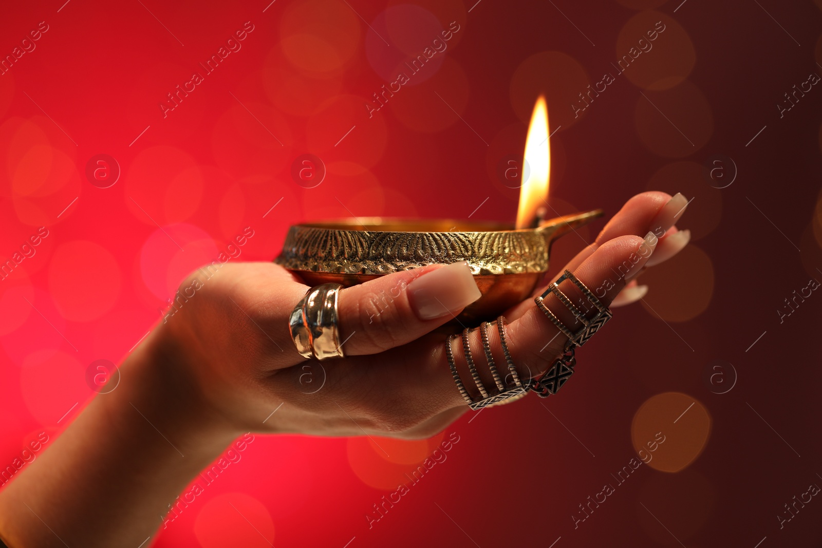 Photo of Diwali celebration. Woman holding lit diya lamp on red background with blurred lights, closeup