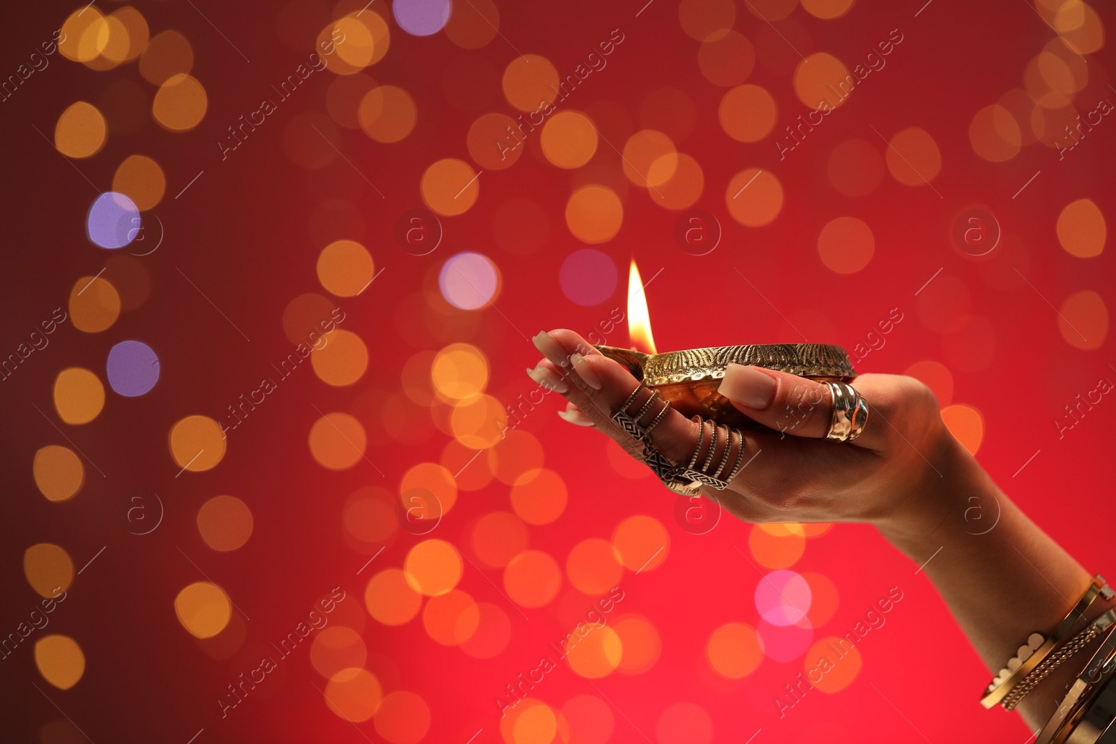 Photo of Diwali celebration. Woman holding lit diya lamp on red background with blurred lights, closeup. Space for text