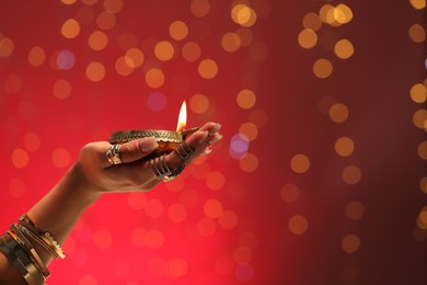 Photo of Diwali celebration. Woman holding lit diya lamp on red background with blurred lights, closeup. Space for text