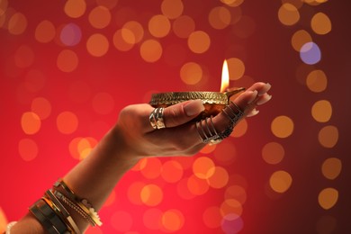 Photo of Diwali celebration. Woman holding lit diya lamp on red background with blurred lights, closeup