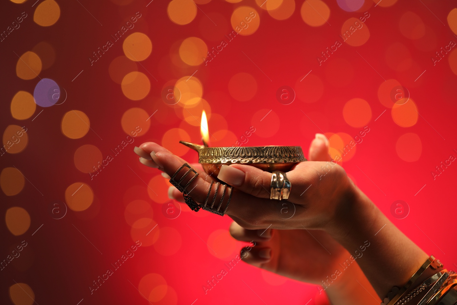 Photo of Diwali celebration. Woman holding lit diya lamp on red background with blurred lights, closeup