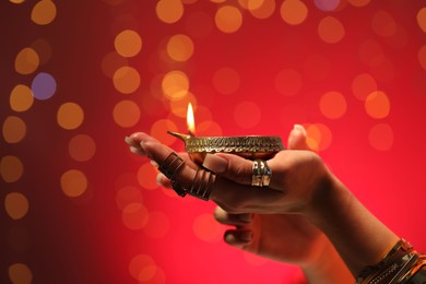 Photo of Diwali celebration. Woman holding lit diya lamp on red background with blurred lights, closeup