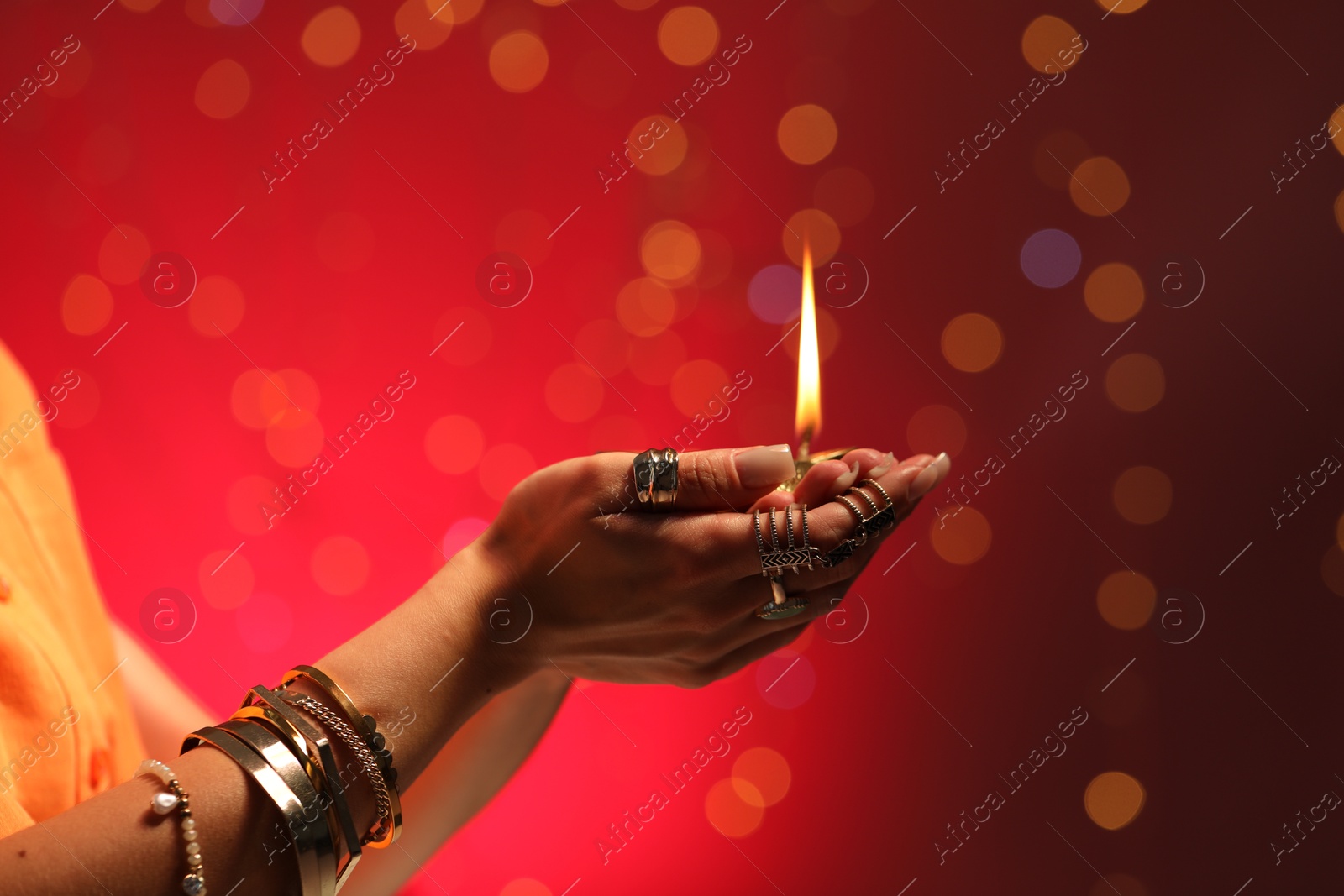 Photo of Diwali celebration. Woman holding lit diya lamp on red background with blurred lights, closeup