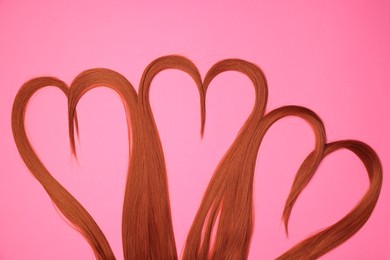 Photo of Red hair strands in shape of hearts on pink background, top view