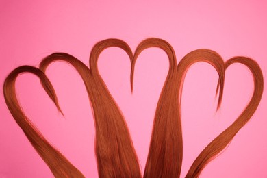 Photo of Red hair strands in shape of hearts on pink background, top view