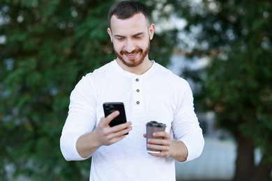 Happy man with paper cup using smartphone on blurred background